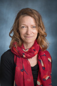 Portrait of a woman with wavy hair wearing a black shirt and a red patterned scarf, smiling against a gradient background.