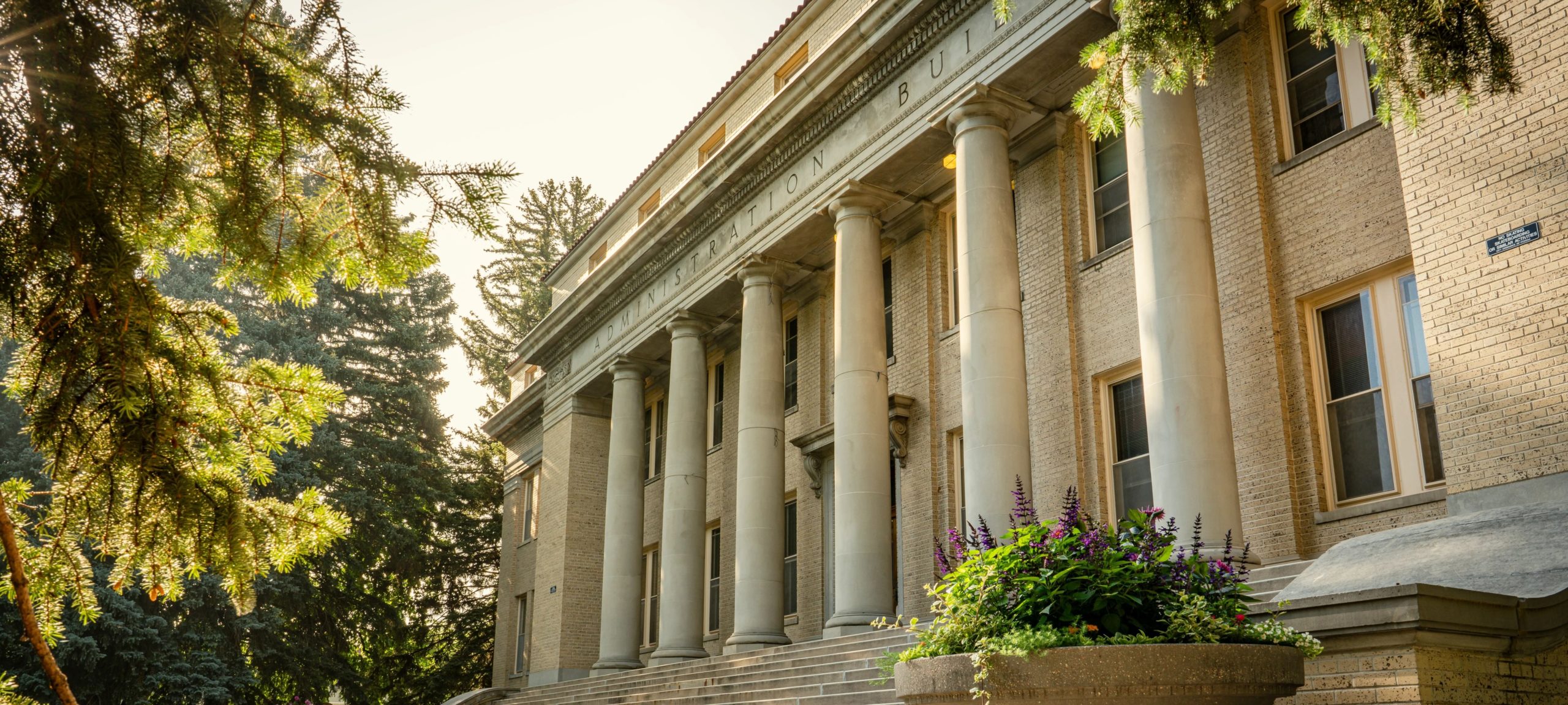 Historic building with large columns, surrounded by greenery, bathed in warm sunlight.