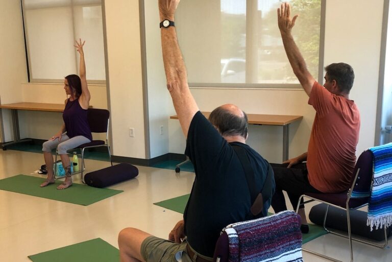 Participants in a seated yoga class raise their hands while practicing mindfulness and stretching in a well-lit room.
