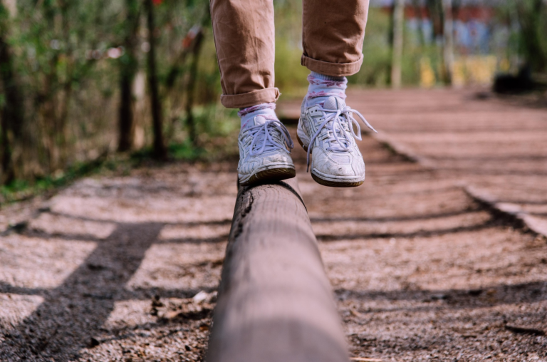 Child balancing on a wooden beam while wearing white sneakers and colorful socks in a natural setting.