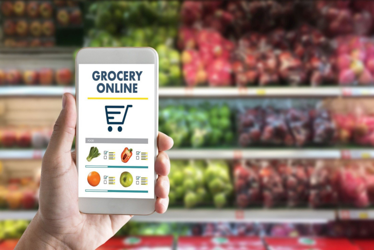 A hand holding a smartphone displaying an online grocery shopping app, with colorful produce in the background.
